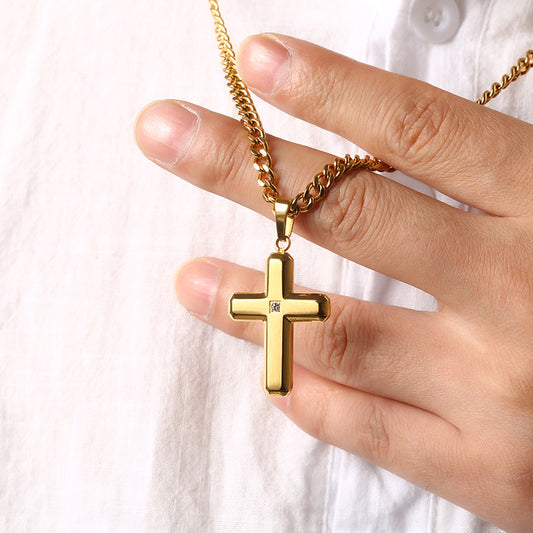A gold cross pendant necklace with a small clear gemstone, held by a hand against a white button-down shirt background.