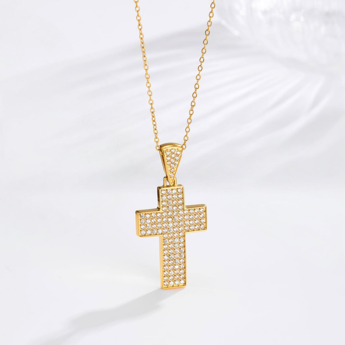 A gold cross pendant necklace paved with small clear stones, hanging from a delicate gold chain against a white background with a soft shadow.