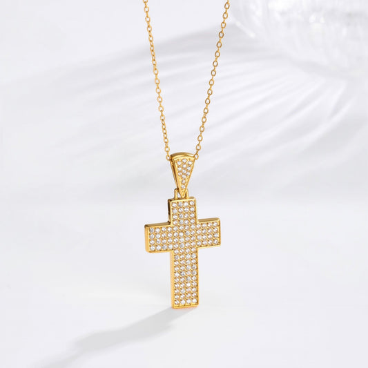 A gold cross pendant necklace paved with small clear stones, hanging from a delicate gold chain against a white background with a soft shadow.