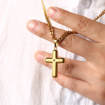 A gold cross pendant necklace with a small clear gemstone, held by a hand against a white button-down shirt background.