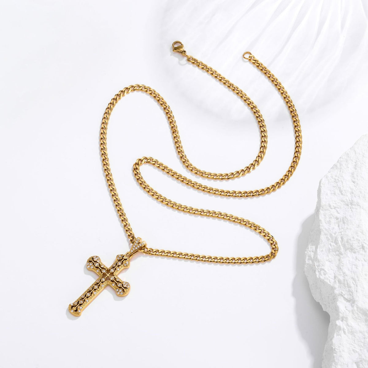 A gold cross pendant necklace with a textured, beaded cross design, displayed on a white background with a white rock prop.